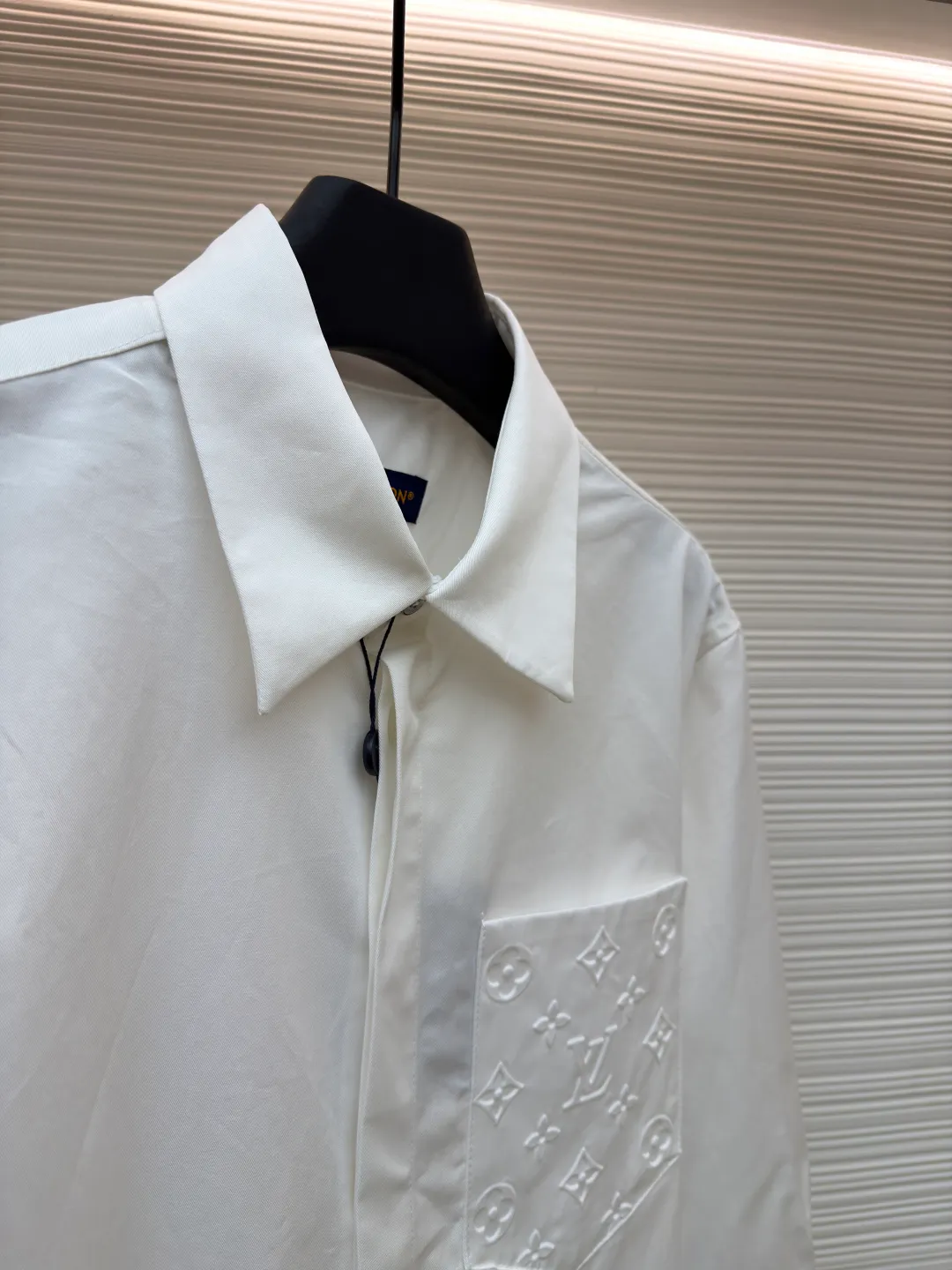 LOUIS VUITTON Embossed Pocket Lapel Shirt ●Original version: Purchased and developed for 3989 ●Item number: 1681491-B05 ●Fabric: Cotton twill woven fabric ●Technology: Concave embossed printing ●Color: white ●Version: Fitted version ●Weight: about 370 grams ●Size: XS-S-M-L ●Description: The twill pattern is obvious on the front of the fabric and blurry on the reverse. Single yarn is used in both warp and weft directions, and the linear density is close. The warp density of the fabric is slightly higher than the weft density, and the hand feel is relatively soft. The fabric has excellent stability, is not easy to deform, and prevents bulging; cotton fiber has good hygroscopicity, which keeps the fabric in a water balance, making people feel comfortable, and adding to the sophistication of the fabric. The concave and convex embossing process is formed by heating and shaping the upper and lower dies. The rollers press the fabric at a certain temperature, and the three-dimensional hierarchical convex surface is produced through mechanical embossing. The fabric produces a concave and convex pattern with a relief-style three-dimensional effect and a special gloss effect. The three-dimensional shape, pattern and text have a strong three-dimensional sense, adding a decorative art effect, clear layers, concentrated three-dimensional image, and accurate perspective angle. The collar can be adjusted with buttons for both formality and comfort; the pleats and tabs at the back are classic shirting techniques, with the pleats providing extra room for movement and the tabs keeping the shirt in shape when hanging. The button front uses custom-fit buttons that are positioned accordingly, and together these details ensure the shirt's functionality and crisp look. The overall wiring is straight and delicate, the fabric is comfortable and soft, and the color is consistent without color difference. It can be matched with the same style to create a fashionable look and control various occasions. Customized accessories, complete with three standards, same style for men and women!
